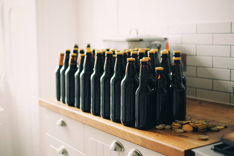 42 brown bottles of beer with yellow caps on a kitchen counter, next to a pile of bottle caps. Home brew heaven.